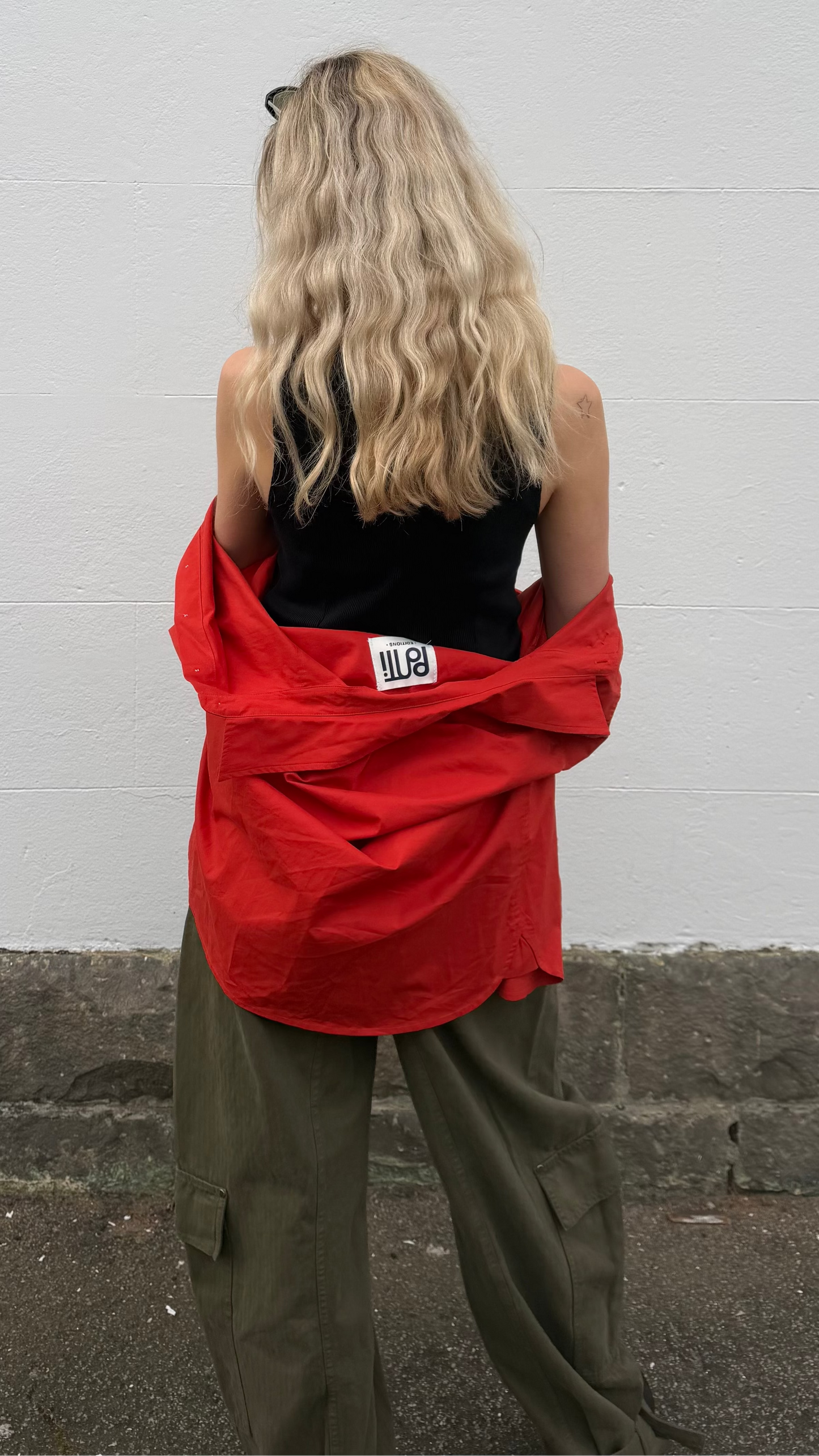 A woman with blonde wavy hair is shown from behind, wearing a bright red oversized button-up shirt tied around her shoulders over a black top and olive green cargo pants, standing against a white painted brick wall.
