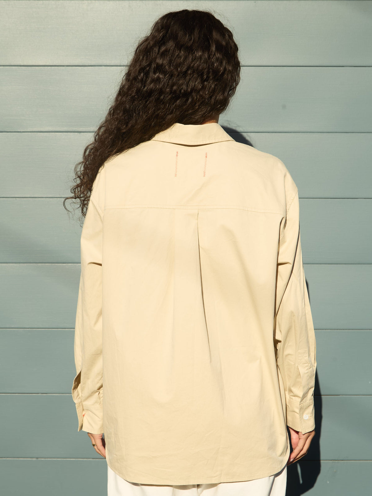 A person viewed from behind wearing a light beige oversized button-up shirt and white pants, standing against a light blue-gray wooden plank wall.